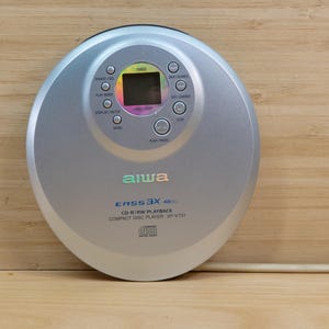 Peut inclure: Lecteur de CD compact Aiwa EASS 3X CD-R/RW argenté. Le lecteur de CD portable a un design circulaire avec un petit écran d'affichage et plusieurs boutons de commande. L'appareil est sur une surface en bois.