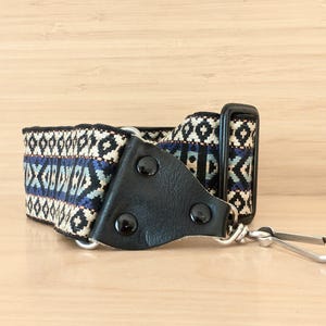 May include: A camera strap with a black leather connector and a patterned fabric strap. The fabric features a geometric design in blue, black, and cream colours. A silver-coloured metal clip is attached to the strap.