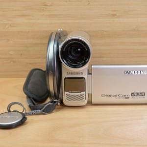 Könnte beinhalten: Silberner Samsung Digital Camcorder mit schwarzem Objektiv und grauem Gurt. Der Camcorder trägt das Samsung-Logo und die Aufschrift "Digital Cam". Der Camcorder liegt auf einer Holzoberfläche.