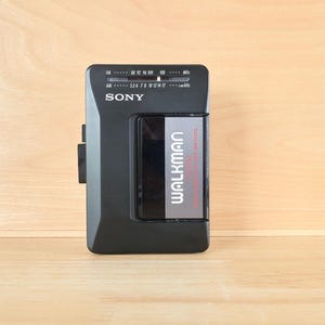 May include: A black Sony Walkman portable cassette player with an integrated AM/FM radio tuner. The device features a rectangular body with a central cassette compartment, a clear window, and white text displaying the Sony and Walkman brand names.