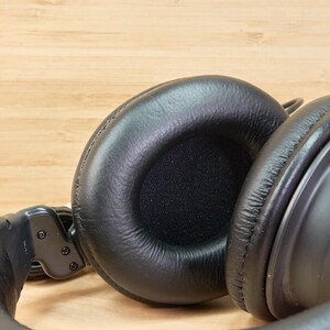 May include: Close-up of black over-ear headphones with large, cushioned earcups. The headphones are made of a smooth, matte material and have a padded headband. The background is a light-colored wooden surface.