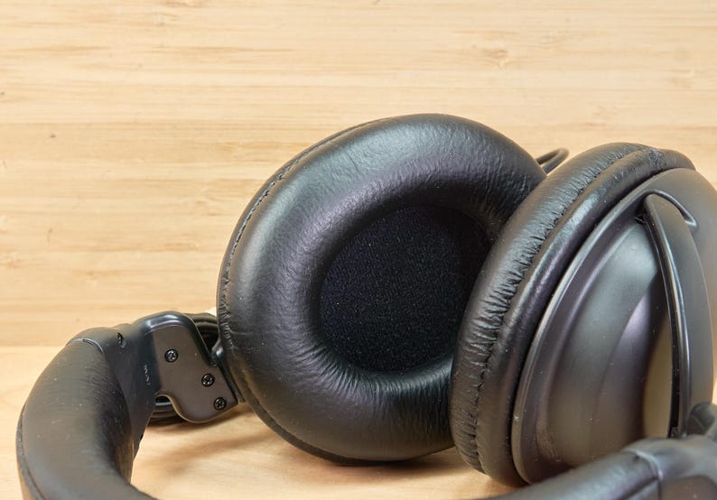 May include: Close-up of black over-ear headphones with large, cushioned earcups. The headphones are made of a smooth, matte material and have a padded headband. The background is a light-colored wooden surface.