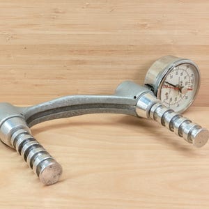 Könnte beinhalten: Ein silbernes Handdynamometer mit einer kreisförmigen Anzeige und zwei geriffelten Griffen. Das Messgerät hat ein weißes Zifferblatt mit schwarzen Markierungen und einen roten Zeiger. Das Gerät dient zur Messung der Griffkraft.