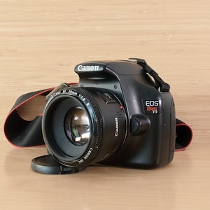 Op de afbeelding: Een zwarte Canon EOS Rebel T3 DSLR-camera met een lensdop en een zwart-rode riem. De lens is een Canon EF 50mm f/1.8. De camera staat op een lichtbruin oppervlak.