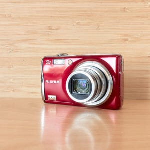 May include: A red Fujifilm digital camera with a silver lens and flash. The camera has a 10x zoom and the brand name is visible. The camera is on a light brown wooden surface.