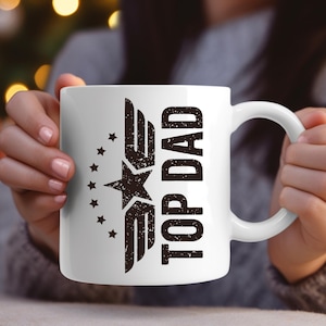 K&ouml;nnte beinhalten: Wei&szlig;e Keramik-Tasse mit dem Text "TOP DAD" in verblichener schwarzer Schrift. Die Tasse hat ein Stern- und Fl&uuml;gel-Design. Die Tasse wird von einer Person gehalten.
