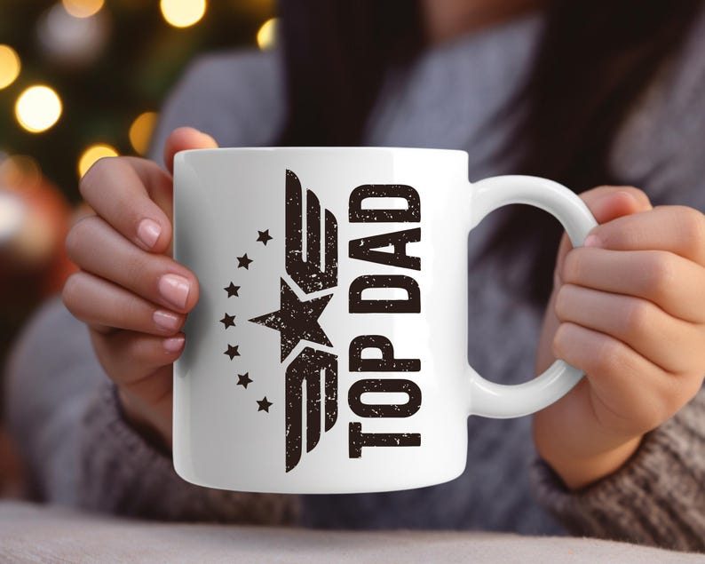 K&ouml;nnte beinhalten: Wei&szlig;e Keramik-Tasse mit dem Text "TOP DAD" in verblichener schwarzer Schrift. Die Tasse hat ein Stern- und Fl&uuml;gel-Design. Die Tasse wird von einer Person gehalten.