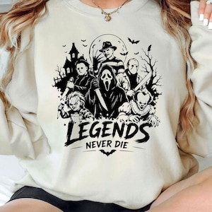 May include: A cream-colored crewneck sweatshirt featuring a black graphic print of various horror movie characters, including a masked figure, a doll, and a cloaked entity. The text "LEGENDS NEVER DIE" is printed in bold black letters below the figures.