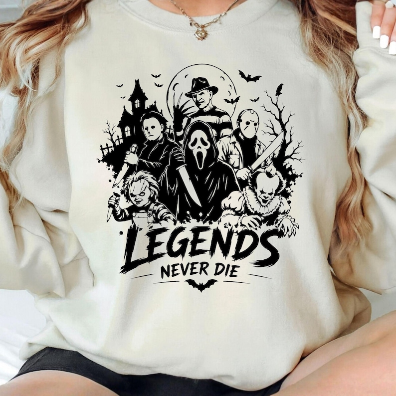 May include: A cream-colored crewneck sweatshirt featuring a black graphic print of various horror movie characters, including a masked figure, a doll, and a cloaked entity. The text "LEGENDS NEVER DIE" is printed in bold black letters below the figures.