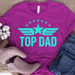 K&ouml;nnte beinhalten: Ein magentafarbenes T-Shirt mit dem Text "TOP DAD" in T&uuml;rkis, mit einem Stern- und Fl&uuml;geldesign. Das Shirt wird mit einer blauen Jeans und einem Paar grauer Stiefeletten kombiniert. Das Design ist im Vintage-Look gehalten.