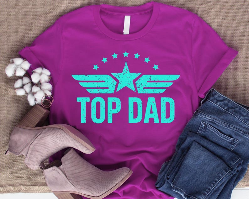 K&ouml;nnte beinhalten: Ein magentafarbenes T-Shirt mit dem Text "TOP DAD" in T&uuml;rkis, mit einem Stern- und Fl&uuml;geldesign. Das Shirt wird mit einer blauen Jeans und einem Paar grauer Stiefeletten kombiniert. Das Design ist im Vintage-Look gehalten.