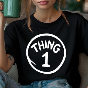 May include: Black t-shirt featuring a white circular graphic with the words "THING 1" in a whimsical font. The number 1 is centered below the word "THING". The shirt is made of a soft material.