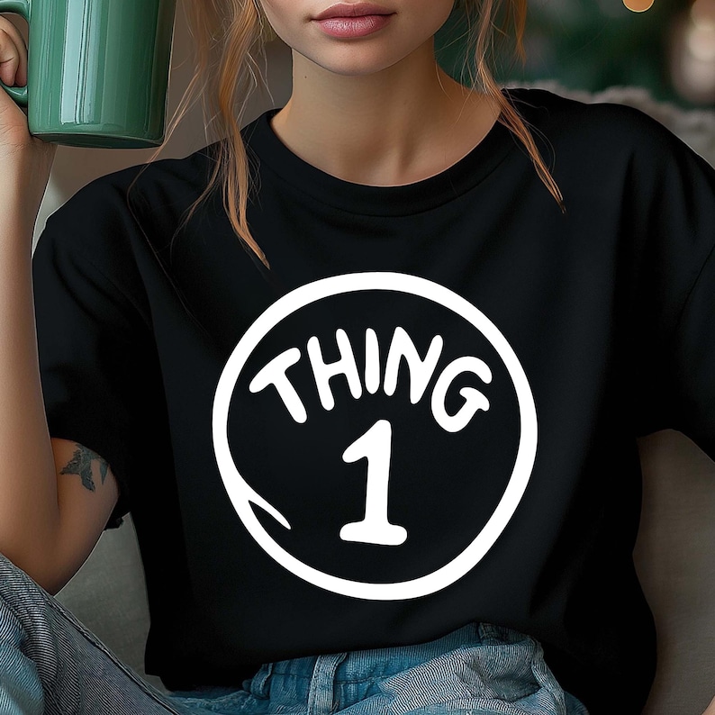 May include: Black t-shirt featuring a white circular graphic with the words "THING 1" in a whimsical font. The number 1 is centered below the word "THING". The shirt is made of a soft material.