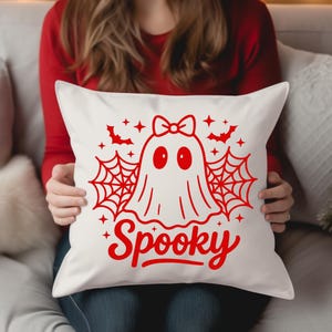 May include: White square pillow with a red Halloween design. The design features a ghost with a bow, spiderwebs, bats, stars, and the word "Spooky" in a script font. The pillow is held by a person wearing a red long-sleeve shirt.