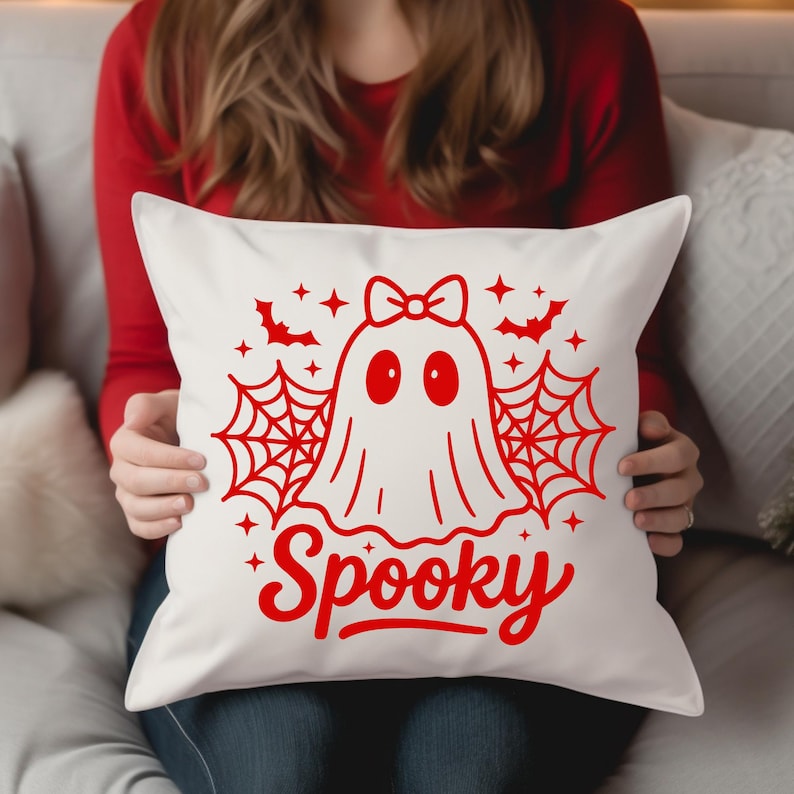 May include: White square pillow with a red Halloween design. The design features a ghost with a bow, spiderwebs, bats, stars, and the word "Spooky" in a script font. The pillow is held by a person wearing a red long-sleeve shirt.