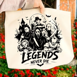 May include: A cream-colored canvas tote bag featuring a black graphic print of five iconic horror movie characters, a haunted house, and bats. Below the characters, the text reads "LEGENDS NEVER DIE" in a bold, stylized font with a small bat icon.