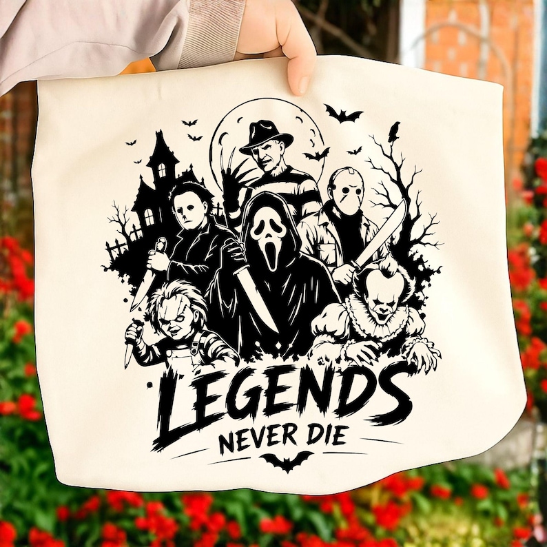 May include: A cream-colored canvas tote bag featuring a black graphic print of five iconic horror movie characters, a haunted house, and bats. Below the characters, the text reads "LEGENDS NEVER DIE" in a bold, stylized font with a small bat icon.