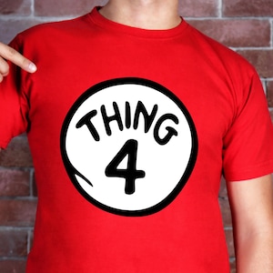 May include: A red t-shirt featuring a white circle with the words "THING 4" in black. The shirt is a solid red color and is being pointed at by a finger.