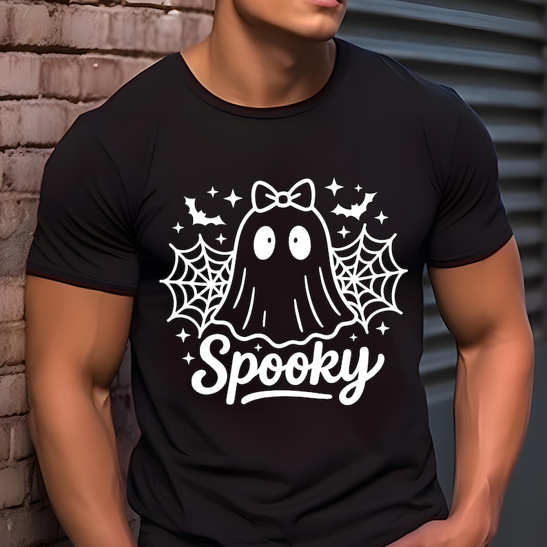 May include: Black t-shirt with a white graphic of a ghost wearing a bow, surrounded by spiderwebs, bats, and stars. The word "Spooky" is written in a script font below the ghost. Halloween-themed apparel.