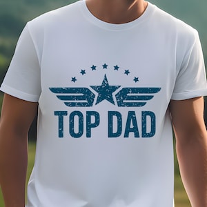 K&ouml;nnte beinhalten: Wei&szlig;es T-Shirt mit blauem Aufdruck "TOP DAD". Das Design beinhaltet einen Stern, Fl&uuml;gel und einen Sternenkranz &uuml;ber dem Text. Das T-Shirt hat kurze &Auml;rmel und scheint aus Baumwolle zu sein.