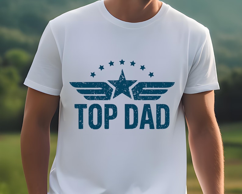 K&ouml;nnte beinhalten: Wei&szlig;es T-Shirt mit blauem Aufdruck "TOP DAD". Das Design beinhaltet einen Stern, Fl&uuml;gel und einen Sternenkranz &uuml;ber dem Text. Das T-Shirt hat kurze &Auml;rmel und scheint aus Baumwolle zu sein.
