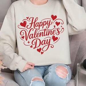 May include: A cream-colored sweatshirt with the text "Happy Valentine's Day" in red script and heart-shaped designs. The design is centered on the front of the sweatshirt. The sweatshirt is a casual, comfortable style.