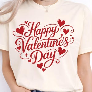 May include: Cream-colored t-shirt featuring a red heart-shaped design with the words "Happy Valentine's Day" in a decorative script. The design includes several small red hearts and flourishes, perfect for Valentine's Day.