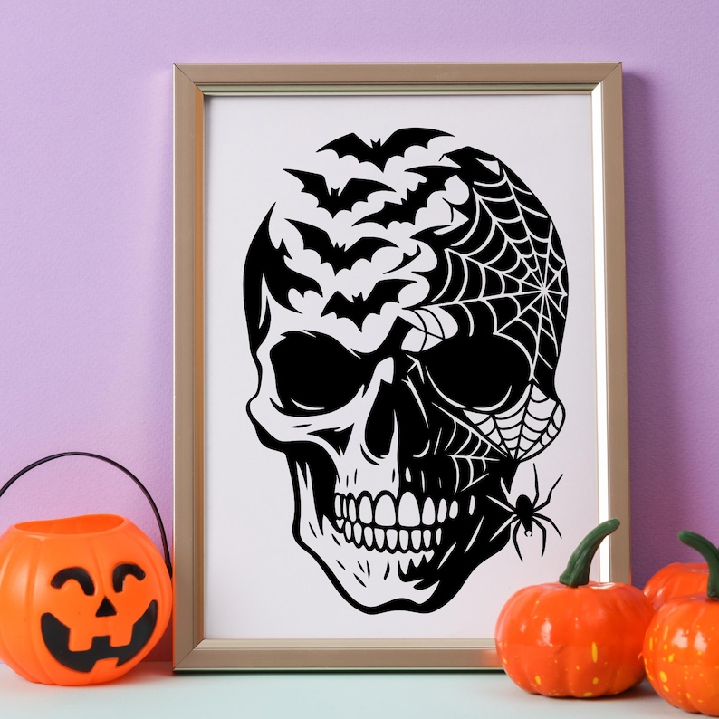 May include: A framed black and white Halloween art print with a skull design. The skull features bats, a spider web, and a spider. An orange jack-o'-lantern bucket and pumpkins are in the foreground.