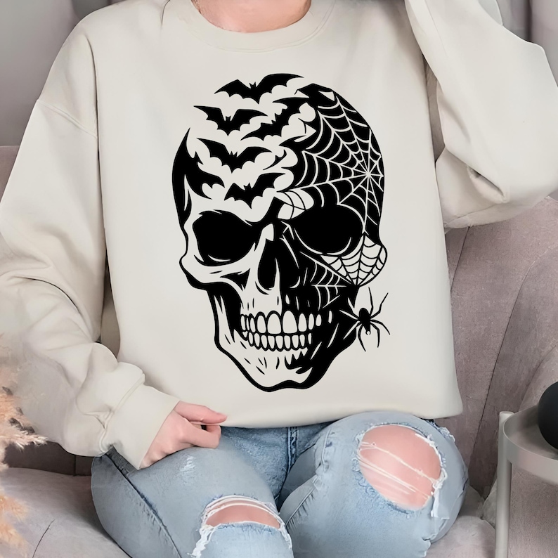 May include: A cream-coloured sweatshirt with a black skull design. The skull is decorated with bats, a spider web, and a spider. The design has a Halloween theme.