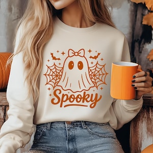 May include: Cream-colored sweatshirt with a Halloween-themed graphic. The design features a ghost with a bow, spiderwebs, bats, and the word "Spooky" in orange. An orange mug is held in the hand.