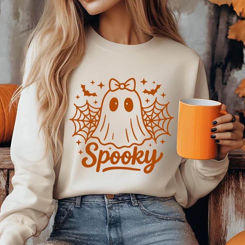 May include: Cream-colored sweatshirt with a Halloween-themed graphic. The design features a ghost with a bow, spiderwebs, bats, and the word "Spooky" in orange. An orange mug is held in the hand.