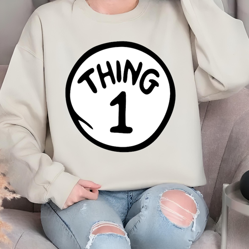 May include: A beige sweatshirt featuring a black and white graphic. The graphic is a white circle with the words "THING 1" in black, with the number 1 in the center. The sweatshirt has long sleeves and a crew neck.