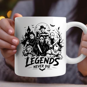 May include: A white ceramic mug featuring a black graphic print of various horror movie characters, including a masked figure, a clown, and a doll, set against a haunted house and moon background with the text "LEGENDS NEVER DIE" at the bottom.
