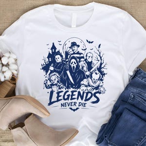 May include: A white short-sleeved t-shirt featuring a dark blue graphic print of various horror movie characters, a haunted house, and bats. The text "LEGENDS NEVER DIE" is printed in a bold, stylized font below the central character illustration.