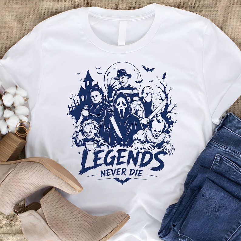 May include: A white short-sleeved t-shirt featuring a dark blue graphic print of various horror movie characters, a haunted house, and bats. The text "LEGENDS NEVER DIE" is printed in a bold, stylized font below the central character illustration.