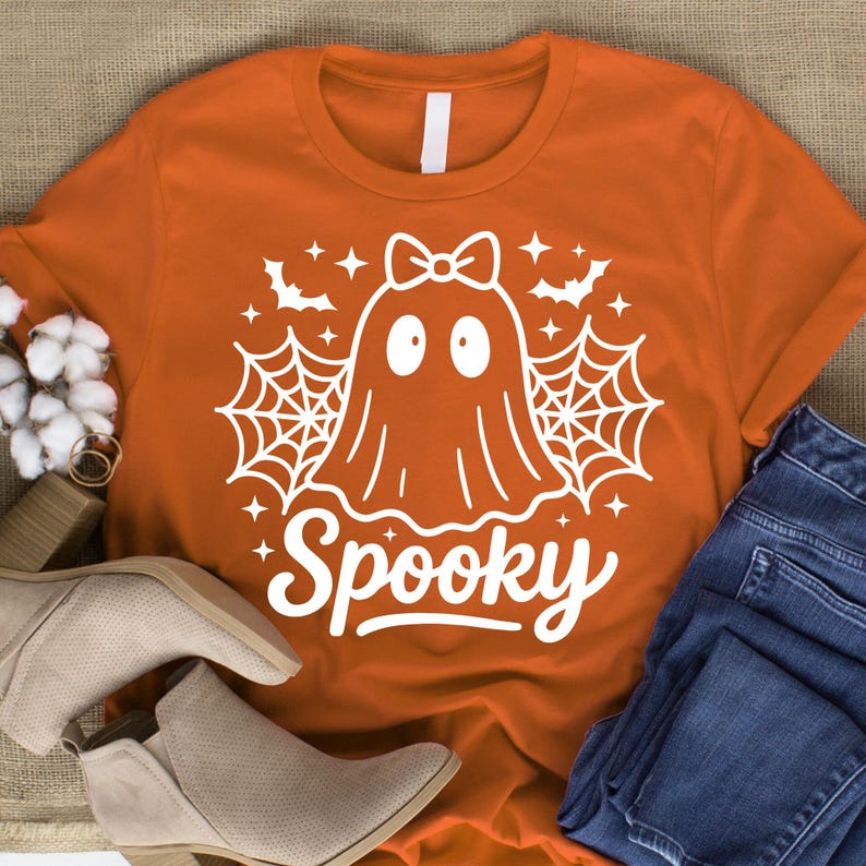 May include: Orange t-shirt with a white ghost graphic, spiderwebs, bats, and the word "Spooky". The ghost has a bow and two oval eyes. The shirt is paired with blue jeans and beige boots.