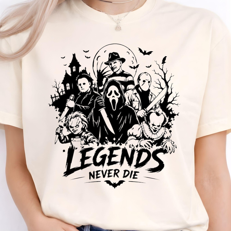 May include: A cream-colored t-shirt featuring a black graphic print of various horror movie characters, including a masked figure, a doll, and a clown. Below the characters, the text reads "LEGENDS NEVER DIE" with a small bat silhouette.