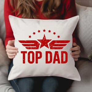 K&ouml;nnte beinhalten: Wei&szlig;es quadratisches Kissen mit roter Grafik "TOP DAD". Die Grafik enth&auml;lt einen Stern, Fl&uuml;gel und kleinere Sterne. Die Person, die das Kissen h&auml;lt, tr&auml;gt ein rotes Langarmshirt.