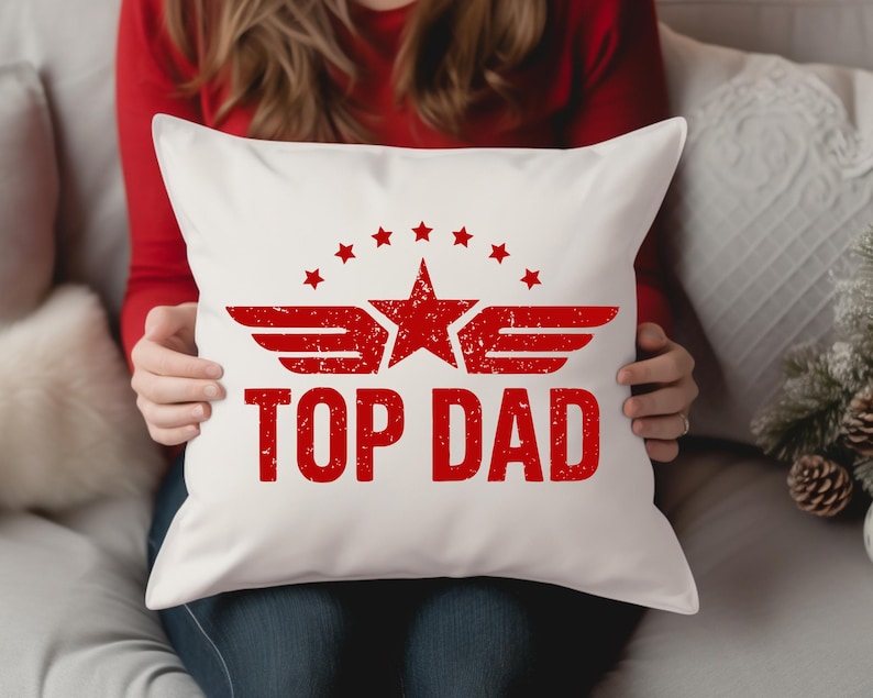 K&ouml;nnte beinhalten: Wei&szlig;es quadratisches Kissen mit roter Grafik "TOP DAD". Die Grafik enth&auml;lt einen Stern, Fl&uuml;gel und kleinere Sterne. Die Person, die das Kissen h&auml;lt, tr&auml;gt ein rotes Langarmshirt.