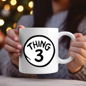 May include: White ceramic mug featuring the text "THING 3" in black within a black outline on a white circle. The mug has a curved handle and is held by a person.