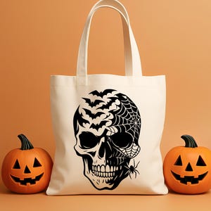 May include: A cream tote bag with a black skull design, bats, a spider web, and a spider. Two carved orange pumpkins are either side of the bag, creating a Halloween theme.
