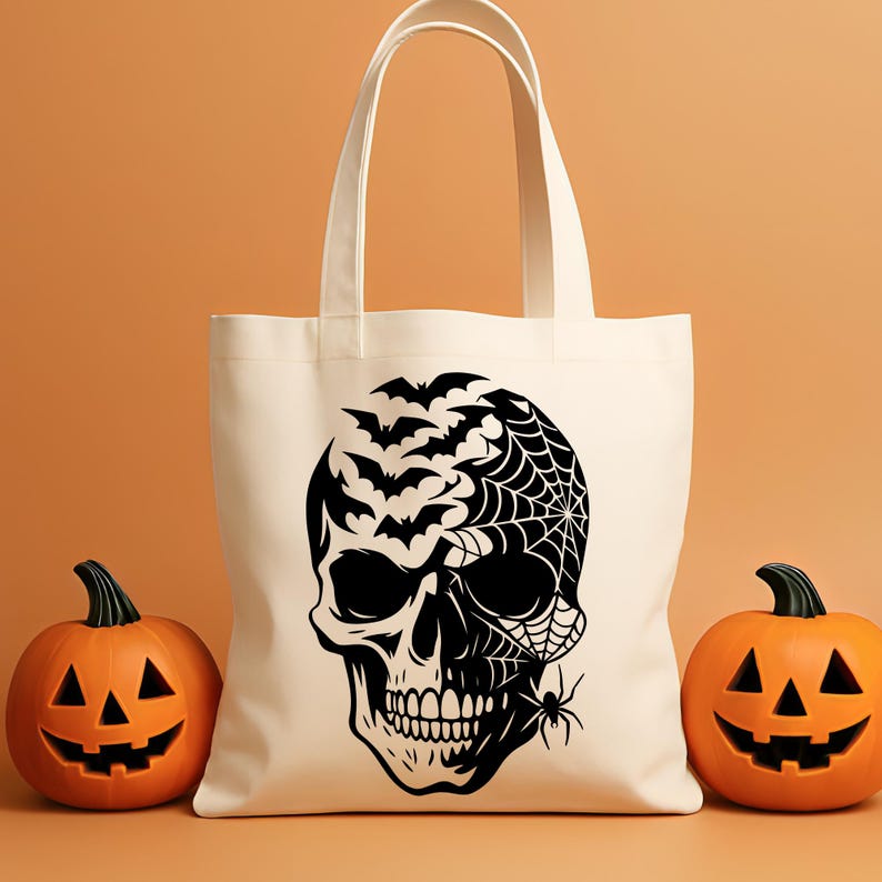 May include: A cream tote bag with a black skull design, bats, a spider web, and a spider. Two carved orange pumpkins are either side of the bag, creating a Halloween theme.
