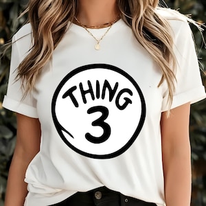 May include: White t-shirt with rolled sleeves featuring a black and white graphic. The graphic is a circle with the words "THING 3" in a playful font. The shirt is a casual style.