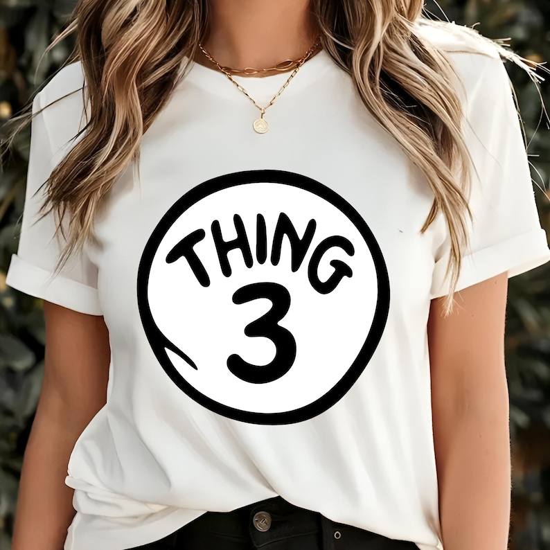 May include: White t-shirt with rolled sleeves featuring a black and white graphic. The graphic is a circle with the words "THING 3" in a playful font. The shirt is a casual style.