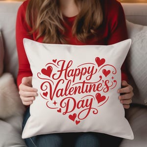 May include: A white square pillow with the words "Happy Valentine's Day" in red script and heart designs. The pillow is held by a person wearing a red long-sleeve shirt.