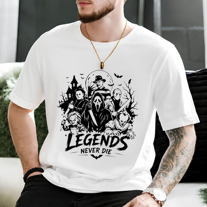 May include: A white short-sleeved t-shirt featuring a black graphic print of various horror movie characters, a haunted house, and bats. Below the characters, the text reads "LEGENDS NEVER DIE" in a bold, stylized font with a small bat icon underneath.