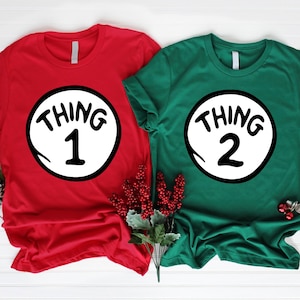 May include: Two short-sleeved t-shirts, one red and one green, each featuring a white circle with black text. The red shirt displays "THING 1" and the green shirt displays "THING 2". The shirts are a festive theme.