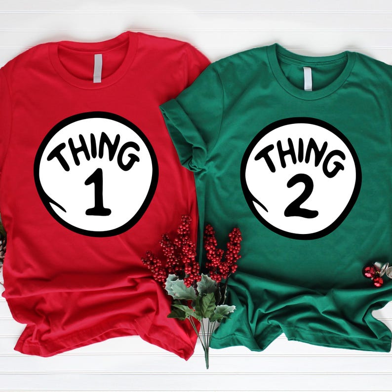 May include: Two short-sleeved t-shirts, one red and one green, each featuring a white circle with black text. The red shirt displays "THING 1" and the green shirt displays "THING 2". The shirts are a festive theme.