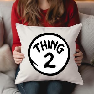 May include: A white square pillow with the words "THING 2" in black, inside a black circle. The pillow is held by a person wearing a red long-sleeve shirt and blue jeans. The pillow is on a white couch.