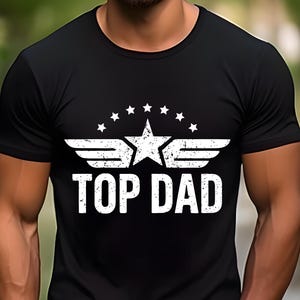 K&ouml;nnte beinhalten: Schwarzes Rundhals-T-Shirt mit einem wei&szlig;en, verwitterten Aufdruck. Der Aufdruck zeigt einen Stern, Fl&uuml;gel und den Text "TOP DAD" in einer fetten, serifenlosen Schrift. Das Design ist auf der Brust zentriert.